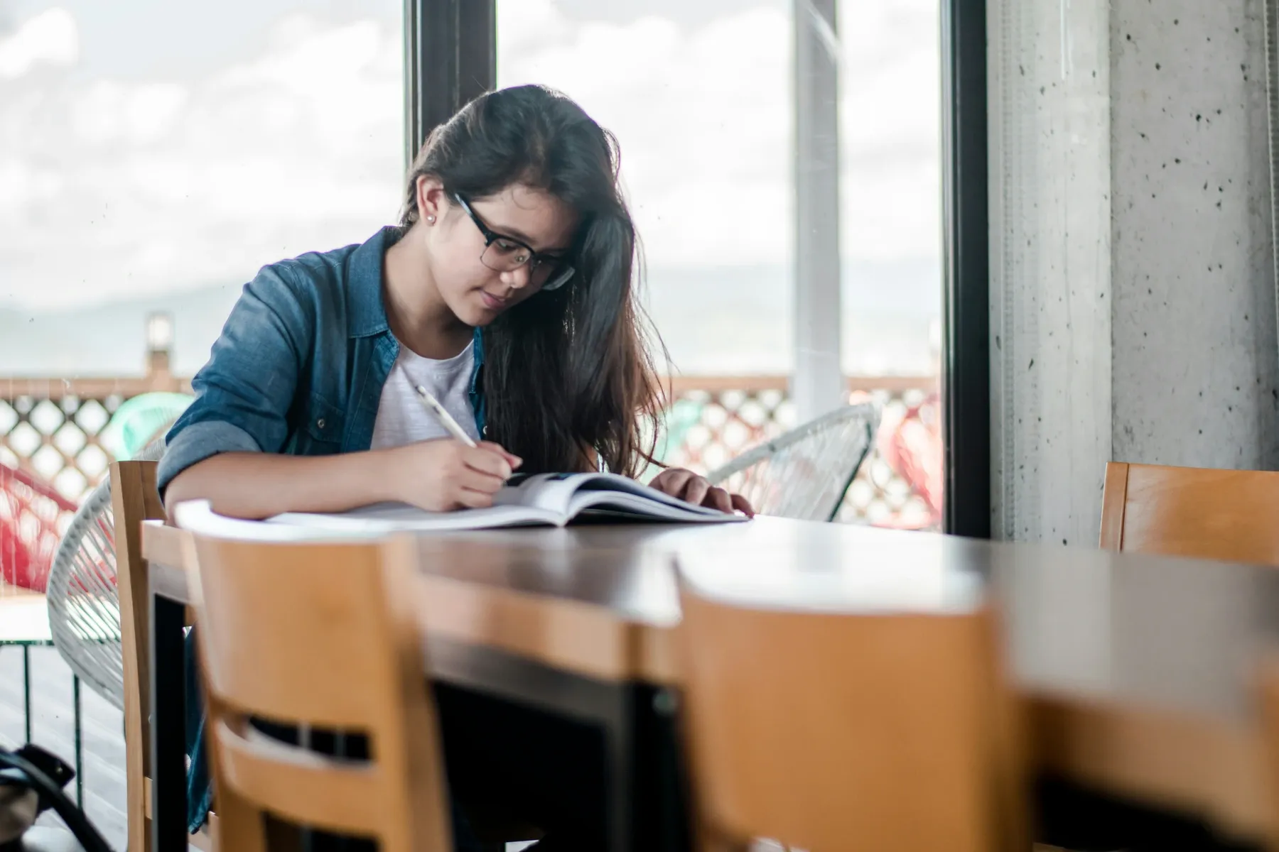 Una joven con gafas toma apuntes junto a un libro abierto en una mesa de estudio.